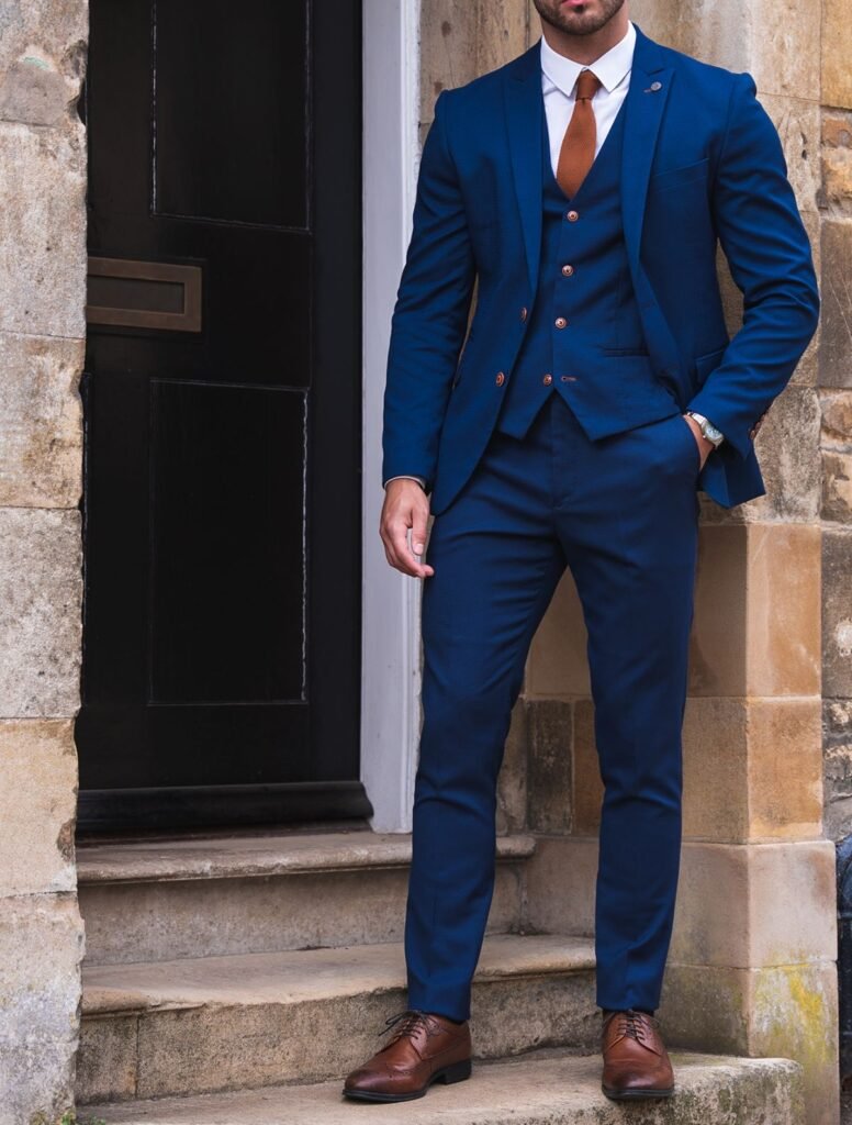 Man in deep navy three‑piece business formals with white shirt, brown tie, and tan Oxford shoes standing by a stone doorway—business formal outfit.