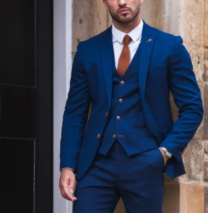Man in deep navy three‑piece business formals with white shirt, brown tie, and tan Oxford shoes standing by a stone doorway—business formal outfit.